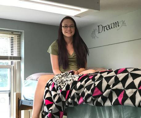 Image alt. text: photo of sophomore Alex on a lofted dorm room bed with a hot pink, black, and white bedspread. She is smiling and looks a little terrified.